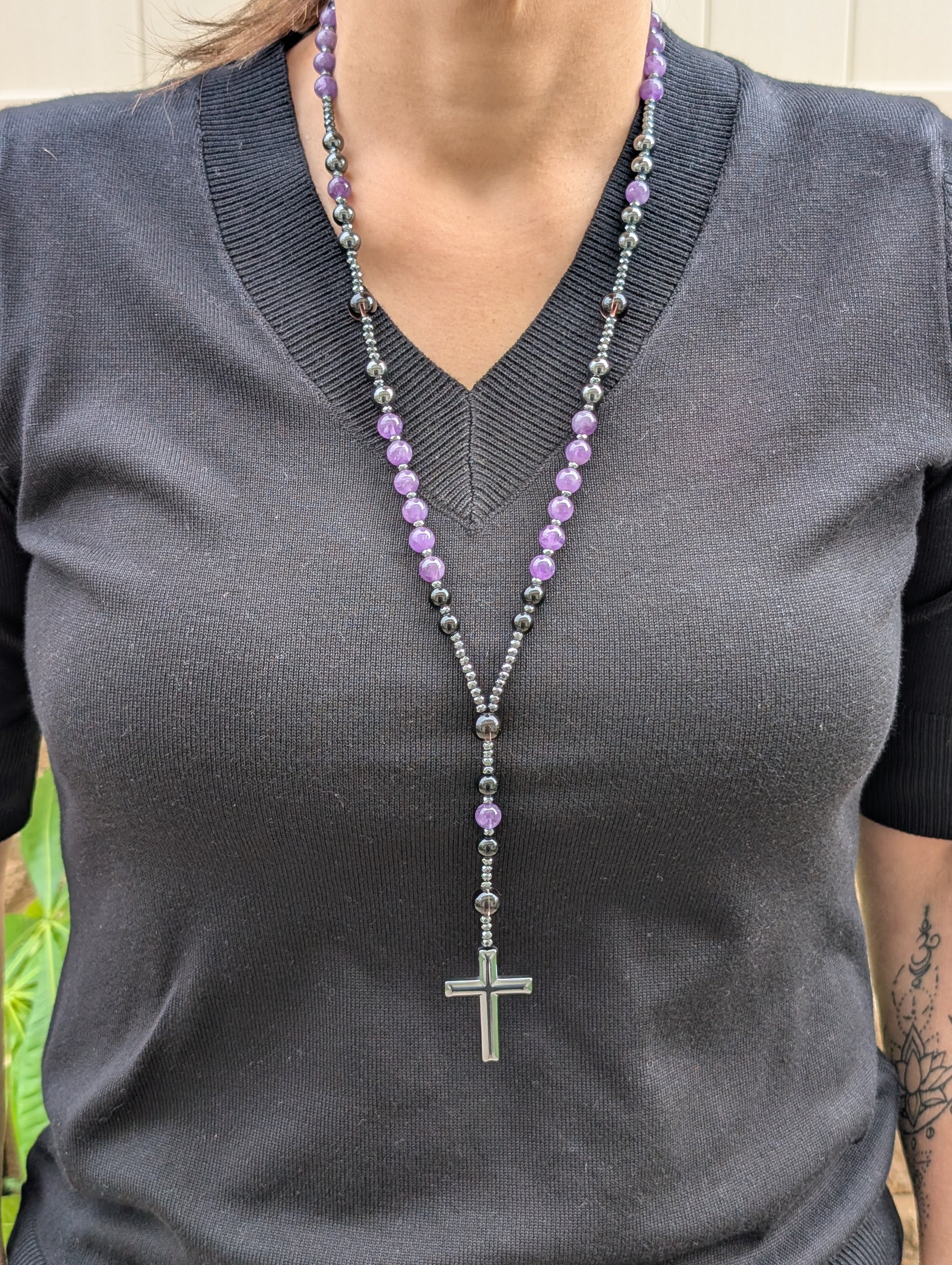 Person wearing a gray shirt with a long beaded necklace featuring purple and black beads and a cross pendant.
