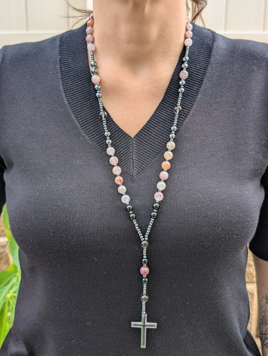 Person wearing a long beaded necklace with a cross pendant on a dark shirt.