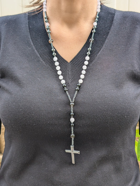 Person wearing a beaded necklace with a cross pendant on a gray shirt.