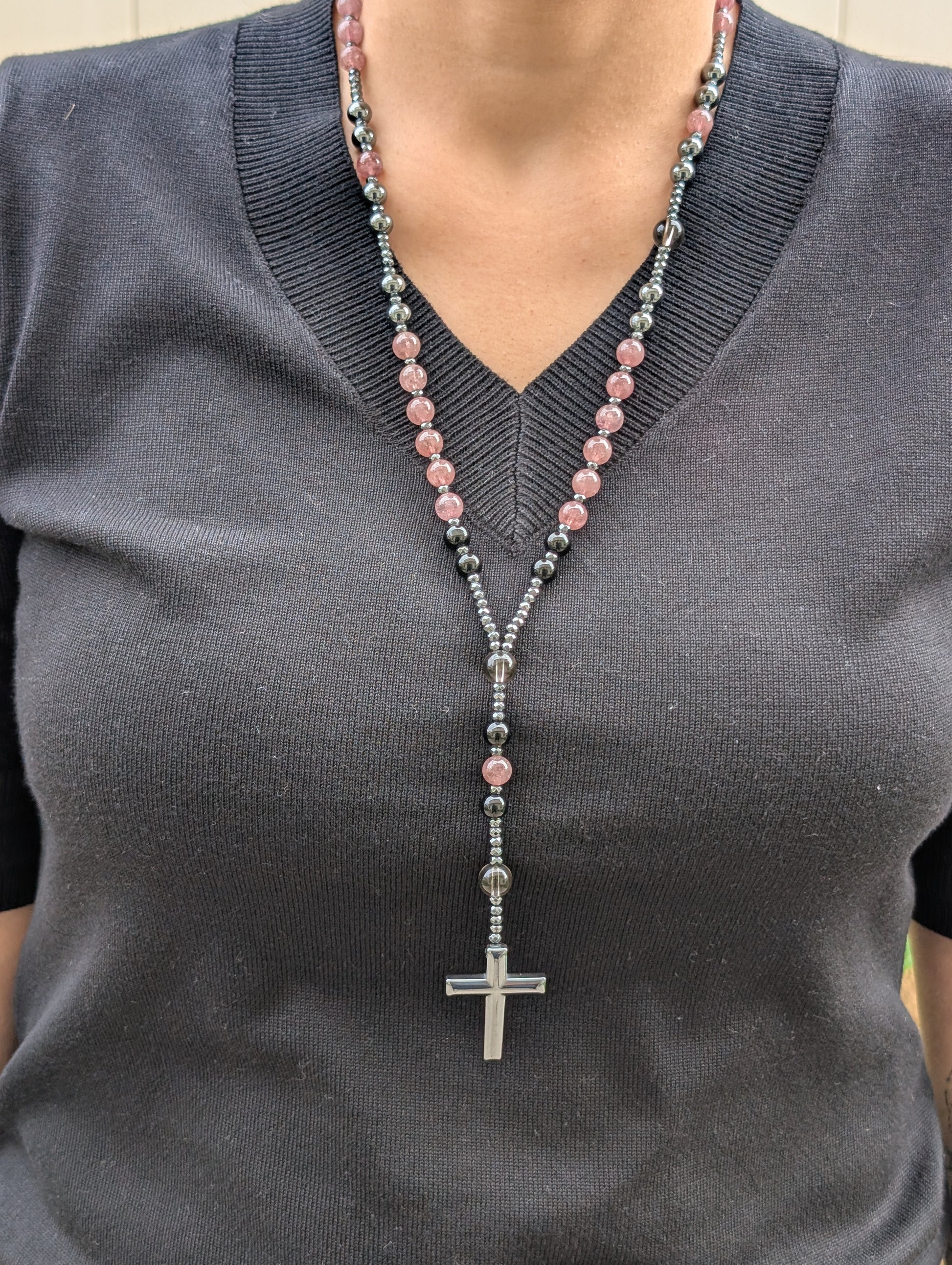 Person wearing a dark gray sweater with a beaded necklace featuring pink and black beads and a silver cross pendant.