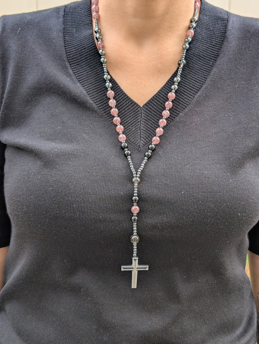 Person wearing a dark gray sweater with a beaded necklace featuring pink and black beads and a silver cross pendant.